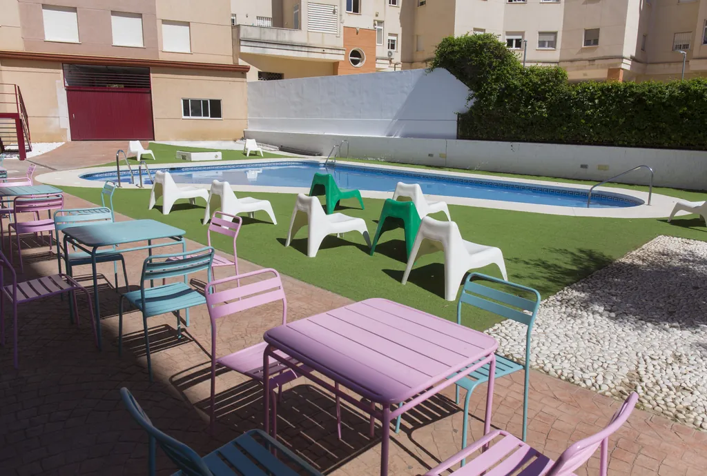 Patio de la residencia con la piscina, césped y mesas de colores al sol